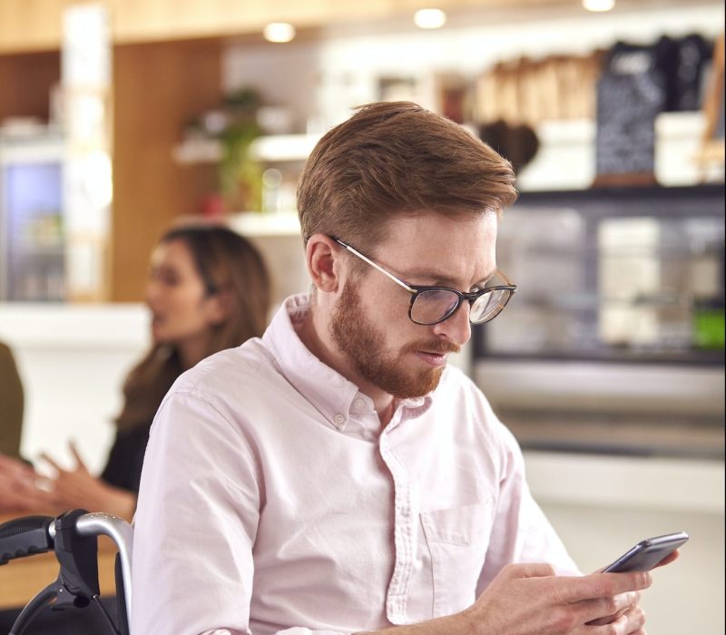 Businessman In Wheelchair With Mobile Phone Sitting At Desk Working On Laptop In Busy Office